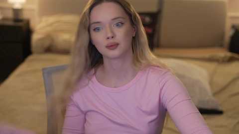 Video of a blonde woman with light skin and blue eyes, wearing a pink long-sleeved top, sitting on a chair in a softly lit bedroom with beige walls and a bed in the background.