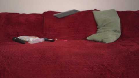 A video of a red velvet couch with a green pillow, a black remote, and a white baby blanket.