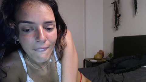 Video of a young woman with dark curly hair, wearing a green tank top, sitting on a bed in a dimly lit room with tribal wall decorations.