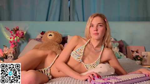 Media: Video of a blonde woman in a crochet bikini, lying on a bed with stuffed toys, surrounded by pastel-colored pillows and a blue curtain.