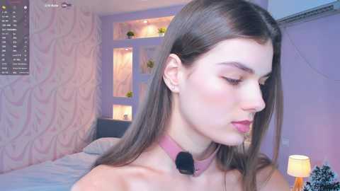 Video of a fair-skinned, brunette woman with straight hair, wearing a pink choker, in a pastel-themed bedroom.