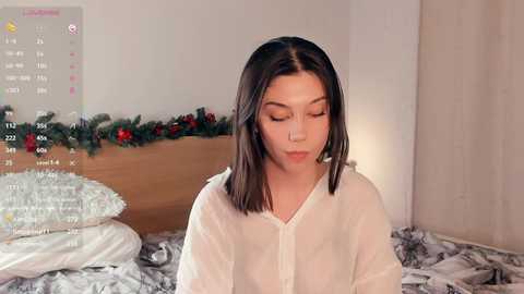 Video of a young woman with fair skin and shoulder-length dark hair, wearing a white button-up shirt, sitting on a bed adorned with holiday decorations and a white duvet.
