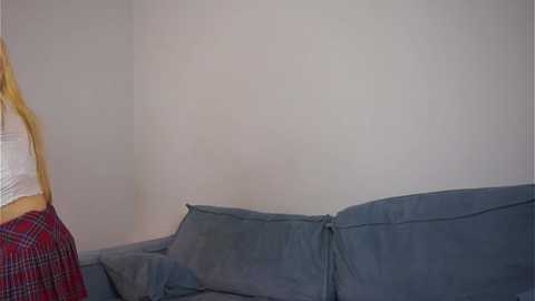 Video of a blonde woman in a white crop top and red plaid skirt, sitting on a grey couch against a plain white wall, capturing a casual indoor scene.