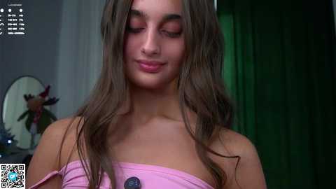 Video of a young woman with long, wet brown hair, wearing a pink off-shoulder top, in a dimly lit room with a green curtain and a round mirror on the left.