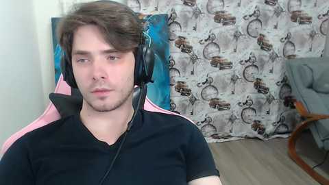 Video of a young man with light skin and brown hair, wearing a black shirt and black headphones, sitting in a pink gaming chair with a blue and brown patterned background.