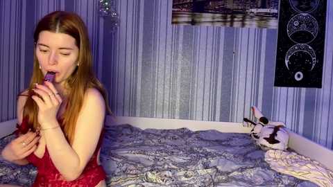 Media: A video of a young woman with long brown hair, wearing a red lace dress, seductively sucking on a purple dildo in a dimly lit, blue-walled bedroom with a cityscape print and a blackboard with circular symbols.