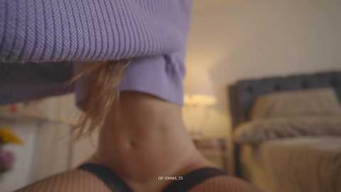 Video of a blonde woman in a purple knit sweater and black bra, her stomach exposed, standing in a dimly lit bedroom with a bed and nightstand in the background.