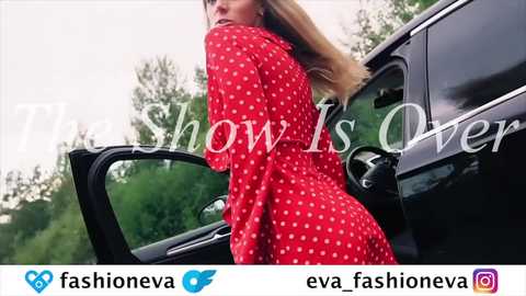 Video of a blonde woman in a red polka-dot dress, leaning into a black car, with green foliage in the background, showcasing her curvaceous figure.