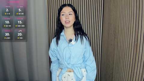 Media: A video of a young Asian woman with long black hair, wearing a light blue button-up shirt tied at the waist, standing in front of a wooden-paneled wall, with a weather forecast display on the left.