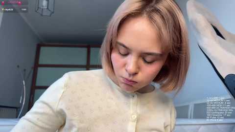 Video of a young Caucasian girl with shoulder-length blonde hair, wearing a cream-colored, textured cardigan, standing indoors, slightly blurred background.
