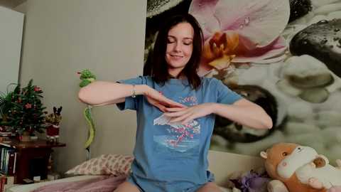 Video of a smiling young woman with shoulder-length brown hair, wearing a blue T-shirt, stretching her arms in a bedroom with a floral wall mural and stuffed animals.
