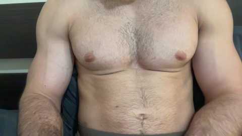 A video of a light-skinned, muscular man with a hairy chest, showing his bare torso and abdomen. He wears dark underwear. Background includes a dark wooden headboard and a blue bedsheet.