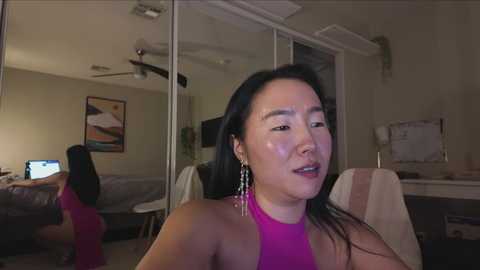 Media: Video of an East Asian woman with long black hair, wearing a bright pink halter top and dangling earrings, sitting in a dimly lit bedroom.