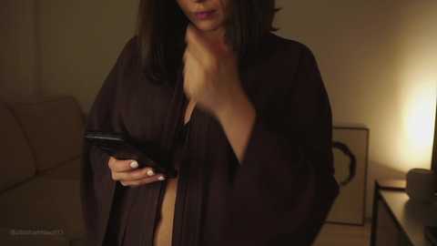 Video of a woman with long, dark hair, wearing a black robe, holding a black smartphone, in a dimly lit living room with beige sofa, lamp, and framed artwork.