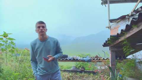 Video of a young man with short dark hair, wearing a blue long-sleeve shirt and gray pants, standing outdoors near a rustic wooden structure with a corrugated metal roof, surrounded by lush greenery and distant foggy mountains.