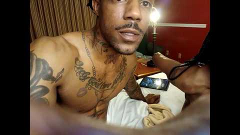 Video of a shirtless, muscular African-American man with tattoos on his chest, posing in a dimly lit room. He has a short beard and is holding a phone.