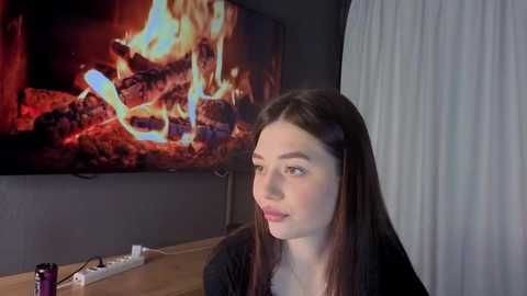 Video of a young woman with long brown hair, fair skin, and light makeup, sitting indoors. She looks to the left, with a TV displaying a burning fireplace in the background.