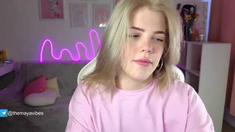 Video of a young, fair-skinned blonde woman with shoulder-length hair, wearing a pink sweater, sitting indoors against a pastel-pink wall with neon \"milk\" lights, pink cushions, and framed artwork.