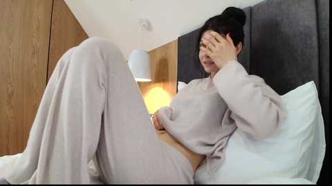 Video of a woman in light gray pajamas, sitting on a bed with a white headboard, covering her face with her left hand, and holding a phone with her right. The room has wooden paneling and a modern pendant light.