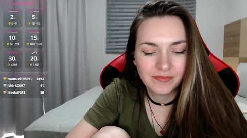 Video of a young woman with long, straight brown hair, wearing a black choker and green top, sitting on a red gaming chair in a modern, minimalistic bedroom with gray curtains and a bed.