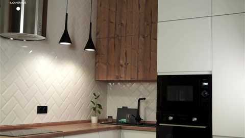 A video of a modern kitchen featuring light-colored cabinets, wooden backsplash, white diamond-patterned tiles, black pendant lights, and a built-in black microwave oven.
