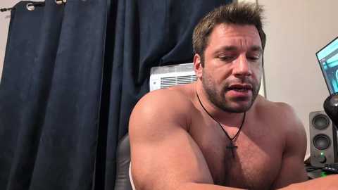 Video of a shirtless, muscular man with short brown hair and a beard, wearing a necklace, looking serious, in a room with dark curtains, a computer, and speakers.