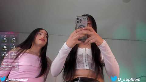 Video of two young women with long black hair, one in a pink top and the other in a white crop top, taking selfies in a dimly lit room with a digital clock display.