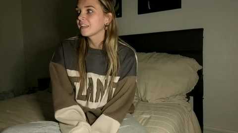 A video of a young woman with long blonde hair, wearing a cozy, oversized sweater, sitting on a bed with white linens in a dimly lit bedroom.