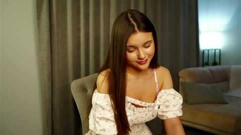 Video of a young, light-skinned woman with long, straight brown hair, wearing a floral off-shoulder dress, sitting on a beige chair in a modern, dimly lit room with gray curtains and a white couch.