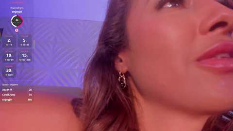 Video of a close-up of a woman with smooth, tanned skin and full lips, wearing dangling black earrings. Background shows a blurred, geometric-patterned wall with purple and blue lighting.