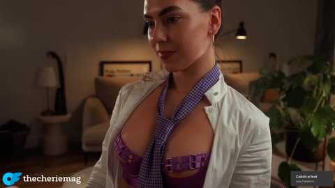 Video of a woman in a purple bra, unbuttoned white shirt, and checkered tie, in a dimly lit living room with potted plants and framed art on the wall.