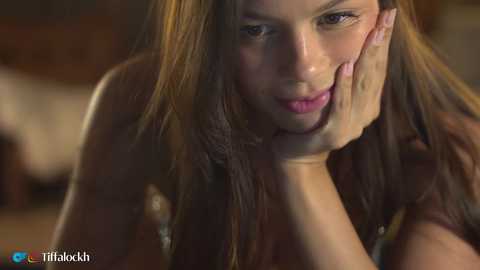 Video of a young woman with long brown hair, fair skin, and pink lips, leaning forward, touching her cheek. She wears a sleeveless top. Background is blurry.