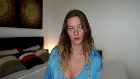 A video of a fair-skinned, slender woman with long, wavy, brown hair. She wears a light blue button-down shirt, revealing a necklace, and stands in a modern bedroom with a bed and wall art.