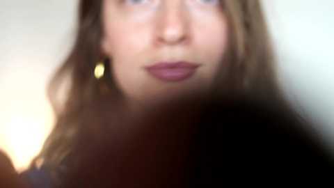 A blurred, close-up video of a young woman with light skin, brown hair, and dark lipstick. She's wearing small gold earrings and has a neutral expression. The background is out of focus, creating a soft, dreamy effect.
