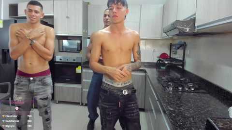 Video of three shirtless men in a modern kitchen. One man with tattoos and a lean physique, wearing a belt, stands in the foreground, while two others, one with a red waistband, are in the background.