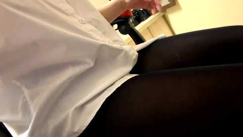 Video of a person in a white blouse, seated, with black tights, using a black office chair with a red armrest. Background includes a beige wall and a desk.