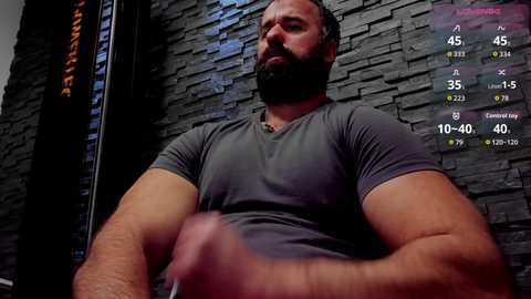 Video of a muscular, bearded man in a gray T-shirt, seated against a dark stone wall. Virtual fitness game display on the right.