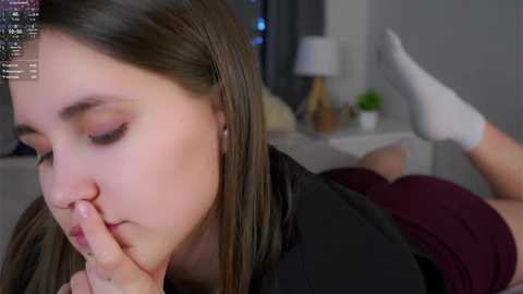 Video of a young Caucasian woman with straight, dark brown hair, lying on her stomach in a dimly lit bedroom, wearing a black top and maroon shorts, with white socks, and gently touching her nose with her finger.