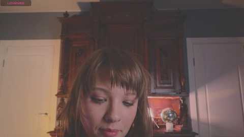 Video of a young woman with light skin and brown hair, wearing a pink top, looking down, in a dimly lit room with wooden furniture and a globe.