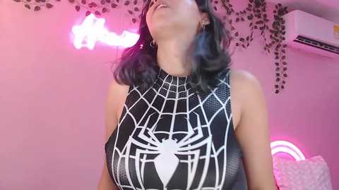 Video of a woman with shoulder-length, wavy dark hair, wearing a black Spider-Man dress with a white spider emblem, against a pink backdrop with hanging vines and a neon sign.