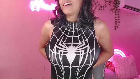 Video of a woman with shoulder-length, wavy black hair, wearing a tight, black Spider-Man costume with a white spider emblem, in a pink-lit bedroom with air conditioning and hanging plants.
