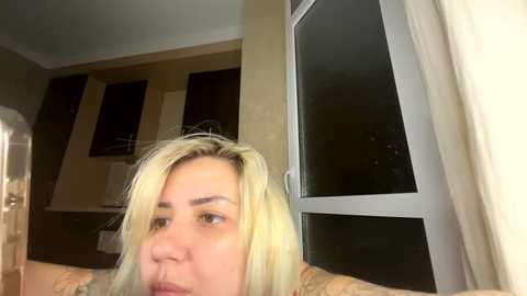 A video of a blonde woman with a tattooed arm, looking out a window with white curtains, in a dimly lit room.