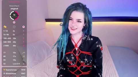 Video of a young woman with pale skin and blue-dyed hair, wearing a black kimono with red rope bondage patterns, sitting on a bed in a softly lit bedroom.