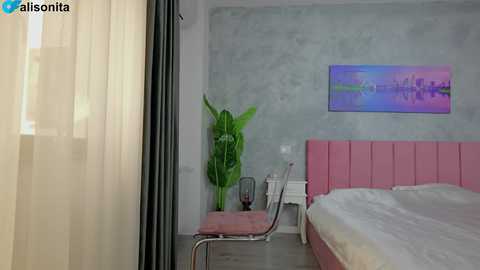 A video of a modern, minimalist bedroom with light gray walls, a pink upholstered headboard, a white bed, a green plant, and a pink side table.