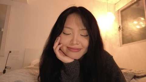 Video of a young Asian woman with long black hair, smiling gently, resting her chin on her hand in a cozy, warmly lit bedroom with a bedside lamp.
