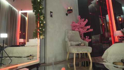 Media: Video of a chic, modern bedroom with a large mirror, plush cream armchair, and floral decor, lit by warm red lights.