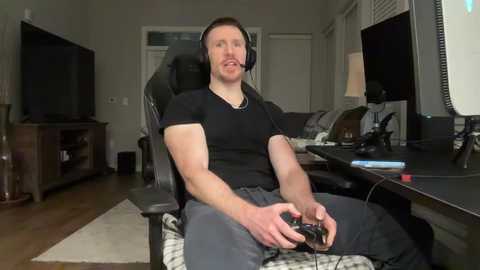 Video of a muscular man with short brown hair, wearing a black shirt and gray pants, sitting in a black chair, gaming on a PlayStation controller in a modern, cluttered living room.