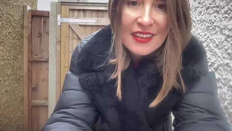 Video of a smiling woman with light skin, straight brown hair, and red lipstick, wearing a black puffer jacket, standing in front of a wooden gate and textured wall.