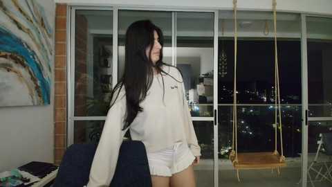 Video of a young woman with long black hair, wearing a white t-shirt and shorts, standing by a window overlooking a cityscape at night.