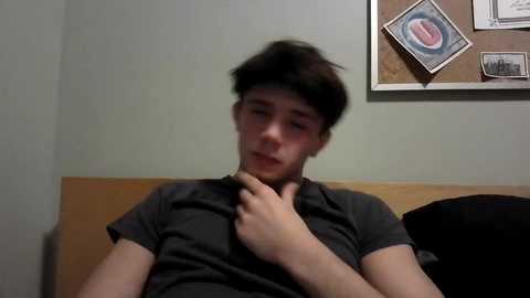 A video of a young man with short brown hair, wearing a dark T-shirt, sitting on a wooden bed with a light-colored wall and a corkboard with framed pictures in the background.